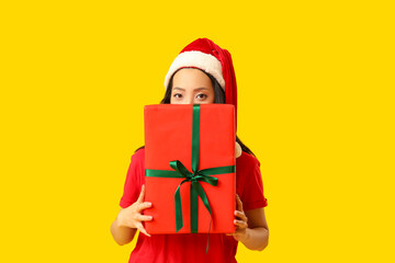 Young Asian woman in Santa hat with Christmas gift box on yellow background