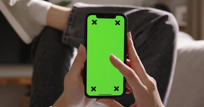 Young woman holds phone with green screen while lounging on sofa, vertical orientation. Advertising app close-up. Tap in center to immersed in captivating video content. World of endless possibilities