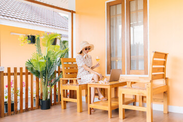Asian female tourists come on holiday to the resort. She sat on a chair in the front of the room. Work with a laptop, hold a glass of wine, wear a dress, glasses, and a hat and feel relaxed.
