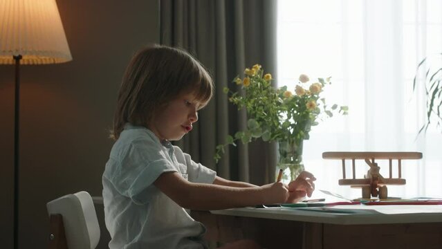 Cute boy draws against background of window at table. Cinematic. Child is passionate about business and creativity. Development of creative potential, happy childhood. People lifestyle, one person