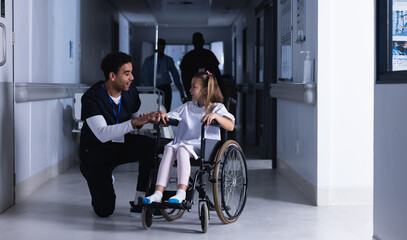 Diverse male doctor talking with girl in wheelchair in hospital corridor
