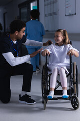 Diverse male doctor talking with girl in wheelchair in hospital corridor