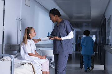 Diverse female doctor talking with girl using tablet in hospital corridor