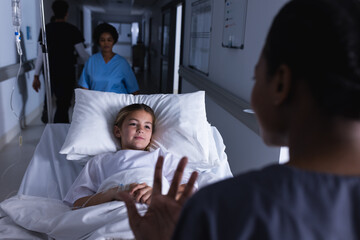 Diverse female doctor talking with girl lying in bed in hospital corridor - obrazy, fototapety, plakaty
