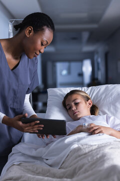 Diverse female doctor using tablet and talking with girl lying in bed in hospital corridor