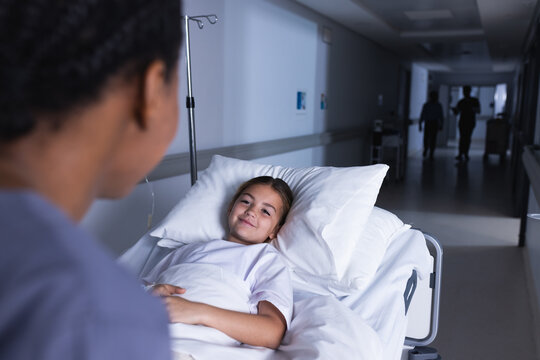 Happy diverse female doctor talking with girl lying in bed in hospital corridor