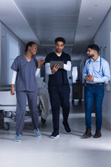 Fototapeta premium Happy diverse male and female doctors using tablet walking in hospital corridor