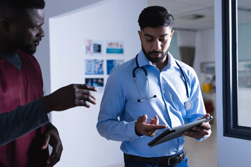 Diverse male doctors talking and using tablet walking in hospital corridor