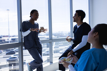 Happy diverse male and female doctors talking, drinking coffee and eating lunch in sunny hospital