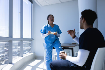 Diverse male and female doctors talking, drinking coffee and eating lunch in sunny hospital