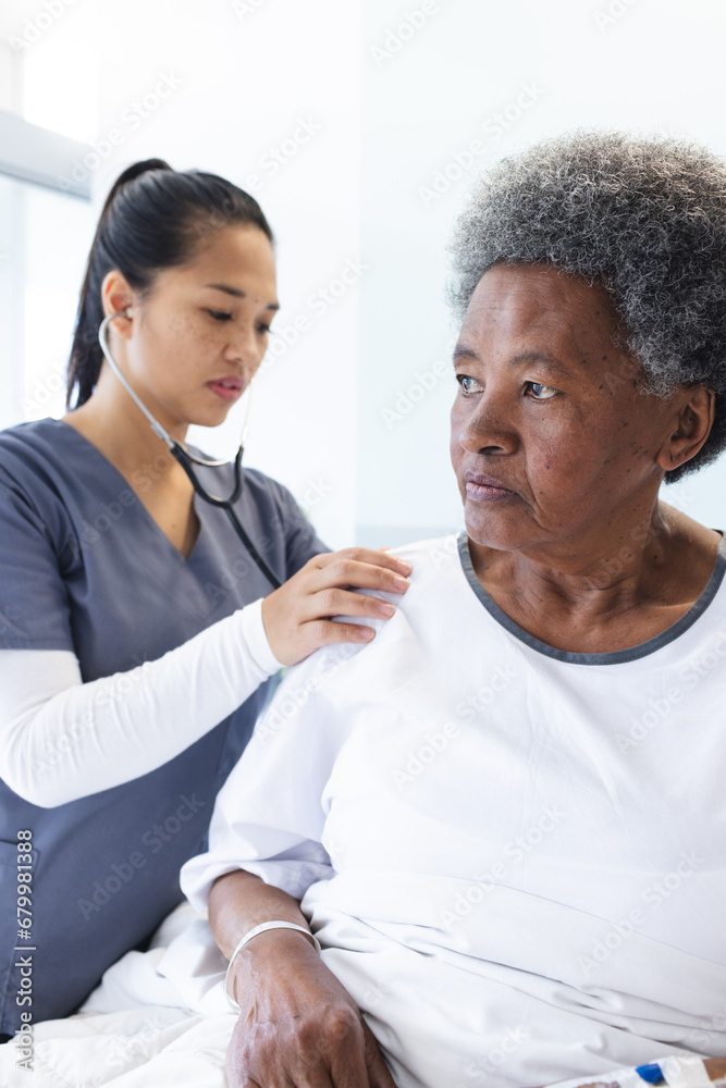 Wall mural Diverse female doctor testing senior female patient using stethoscope in hospital room