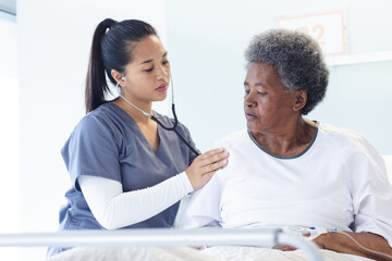 Diverse female doctor testing senior female patient using stethoscope in hospital room