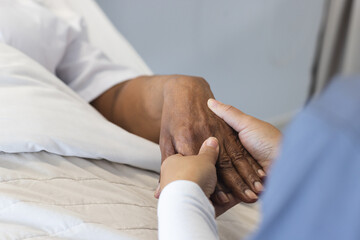 Hands of diverse female doctor holding hand of senior female patient in hospital room