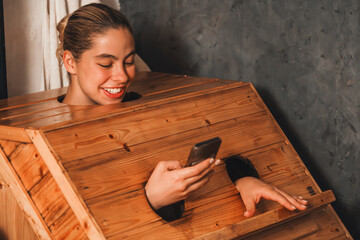 A portrait of gorgeous caucasian woman playing her mobile phone while using wooden sauna cabinet in warm tone. Attractive female with beautiful skin taking a photo. Gray background. Tranquility