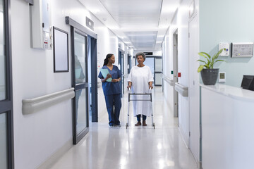 African american female doctor walking with senior female patient with crutches in hospital corridor