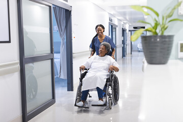 African american female doctor walking with senior female patient in wheelchair in hospital corridor