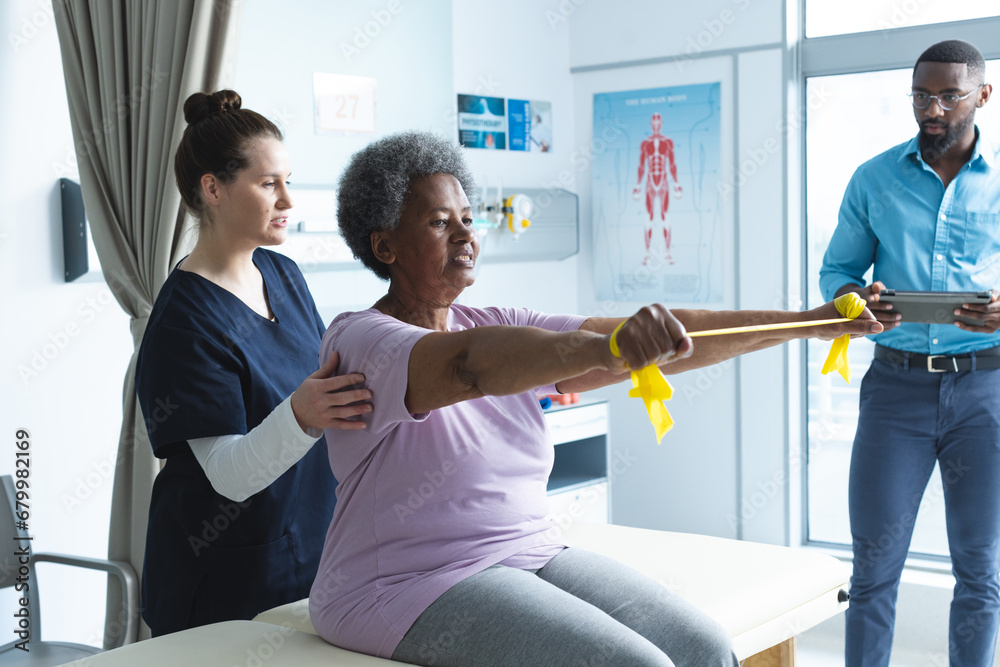 Wall mural Diverse senior female patient exercising with band and female doctor advising in hospital room