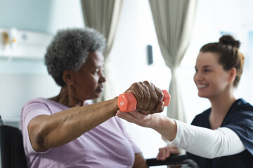 Obraz premium Diverse senior female patient exercising with weight and female doctor advising in hospital room