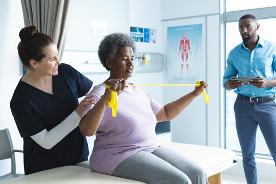 Diverse senior female patient exercising with band and female doctor advising in hospital room