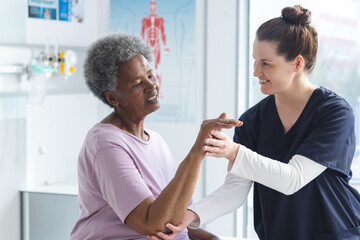 Diverse senior female patient exercising hand and female doctor advising in hospital room