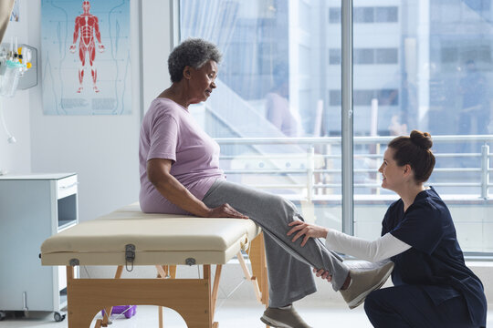 Diverse senior female patient exercising leg and female doctor advising in hospital room