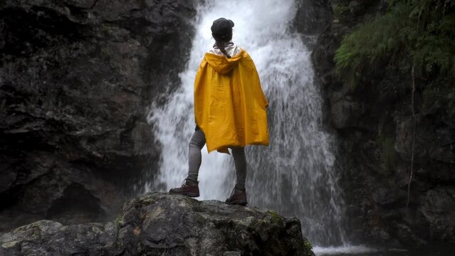 backview of a full lenght silhouette of an asian woman in yellow hooded raincoat and black cap with waterfall on the background