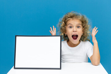 Surprised happy little girl sitting at table with empty white paper with space for text on blue background. Education concept learning. Podium or mockup for advertising. Place for text