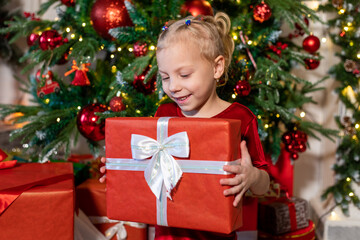 Cute girl with a gift in her hands sits near the Christmas tree.