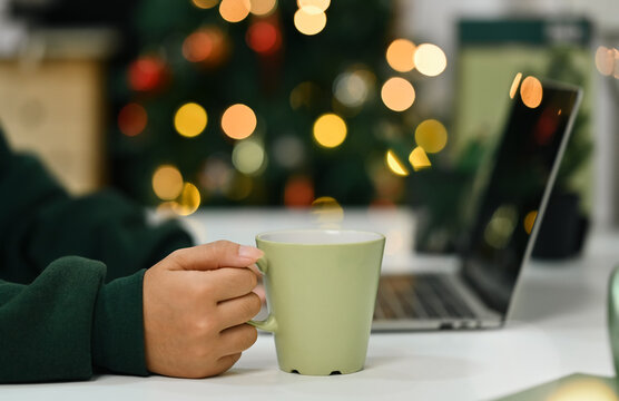 Close up of unrecognizable woman hand holding cup of hot chocolate and using laptop near Christmas tree