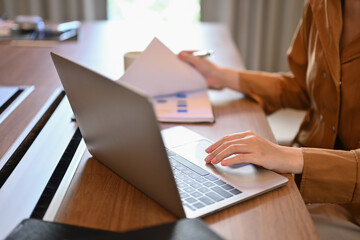 Cropped shot of businesswoman checking reports and using laptop on wooden office desk