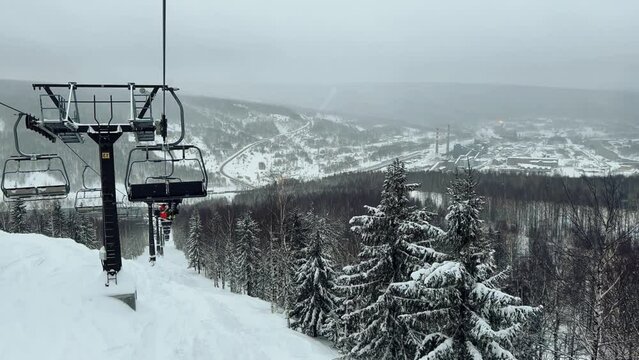 A chairlift at a ski resort on a cloudy winter day in the Ural Mountains. A lift on the background of factory pipes. Climbing the mountain. Gubakha, Perm Krai, 4K