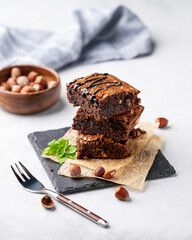 A stack of chocolate brownies with mint, hazelnuts and salted caramel on baking paper on a light background with fork and bowl with nuts.