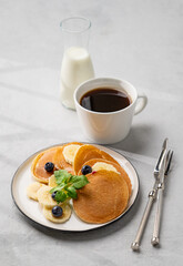 Delicious banana pancakes with fresh fruit slices, berries, mint and honey on a light background with a cup of black coffee, soy milk and shadow.
