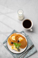 Delicious banana pancakes with fresh fruit slices, berries, mint and honey on a light background with a cup of black coffee, milk and shadow.