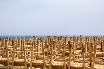 End of season at the seaside concept background. Large group of wooden chairs of closed seafront outdoor cafe or restaurant placed upside down on top of tables, blurry sea horizon in the distance.