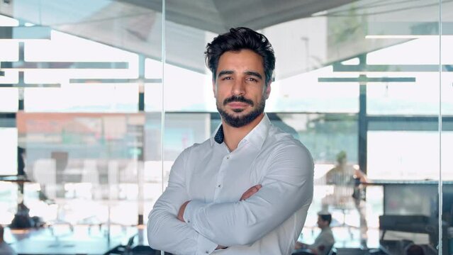 Confident young Latin business man crossing arms standing in office, portrait. Hispanic businessman manager, successful entrepreneur, male professional executive looking at camera.