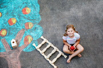 Naklejka premium Cute little girl painting with colorful chalks apples harvest from apple tree on asphalt. Cute preschool child with having fun with chalk picture. Creative leisure for children, drawing and painting.