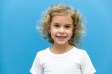 Close up head shot portrait with smiling little blonde curly hair girl. Concept happy and beauty kid with good healthy teeth for dental on blue background, six year child looking at camera and posing - obrazy, fototapety, plakaty