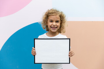 portrait of baby girl holding white drawing Board on multi-colored background, young girl is...