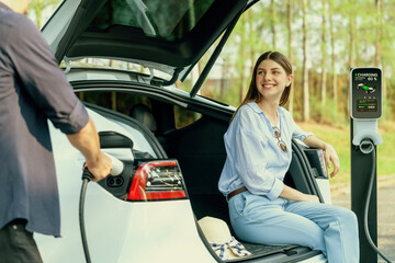 Lovely young couple recharging battery for electric car while relaxing and sitting on the trunk...