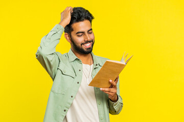 Happy relaxed smiling Indian young man reading funny interesting fairytale story book, leisure hobby, knowledge wisdom, education, learning, study, wow. Arabian Hindu guy isolated on yellow background