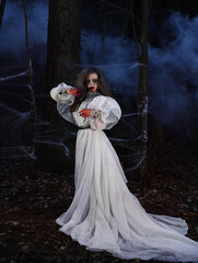 Female ghost in white dress with sickle in hands among smoke in dark forest. Horror photoshoot. Horrid woman with white eyes. Halloween costume, blood on face