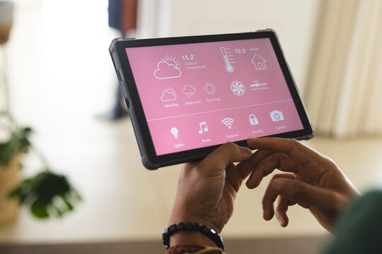 Biracial man with bracelet checking smart home controls on tablet in sunny living room at home