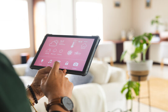 Biracial man with wristwatch checking smart home controls on tablet in sunny living room at home