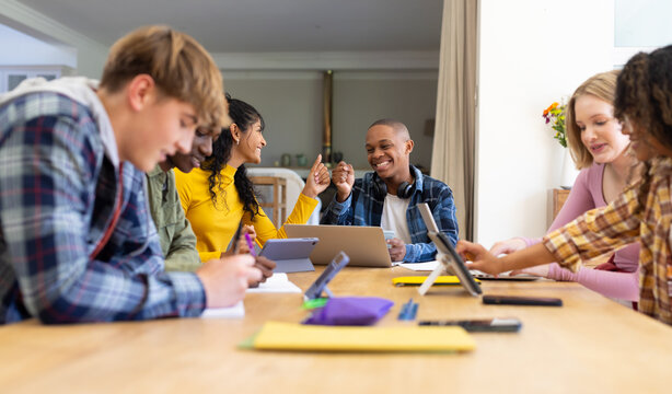 Happy diverse group of teenage friends studying with laptop and tablets at table at home
