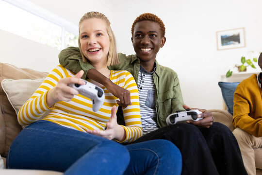 Happy diverse group of teenage friends sitting on couch and playing video games at home