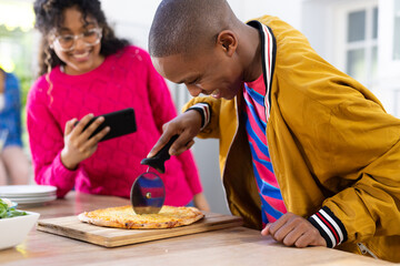 Happy diverse group of teenage friends using smartphone and cutting pizza in kitchen