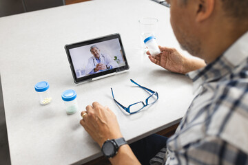 Happy senior biracial man having tablet video call with doctor and taking pills at home