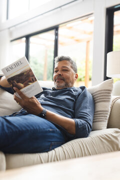 Happy senior biracial man lying on couch and reading book at home