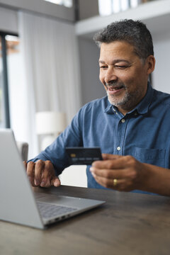 Happy senior biracial man holding credit card and using laptop at home
